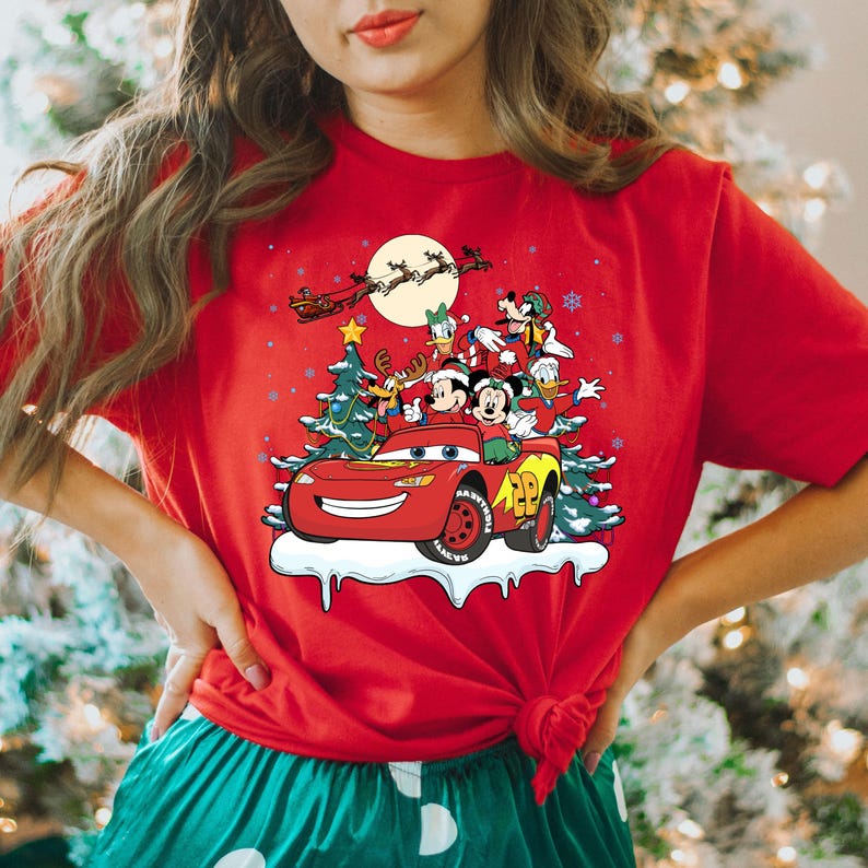 May include: Red t-shirt with a Christmas scene featuring Mickey Mouse, Donald Duck, Goofy, and Pluto riding in a red car with a Christmas tree on top. The car is driving through a snowy scene with a full moon in the background.