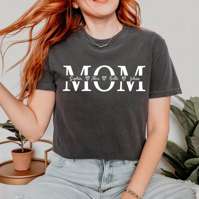 May include: Dark gray t-shirt with the word "MOM" in large white letters. Below the word "MOM" are the names Sophia, Alex, Belle, and Johan, each separated by a heart symbol. The shirt is a casual style.