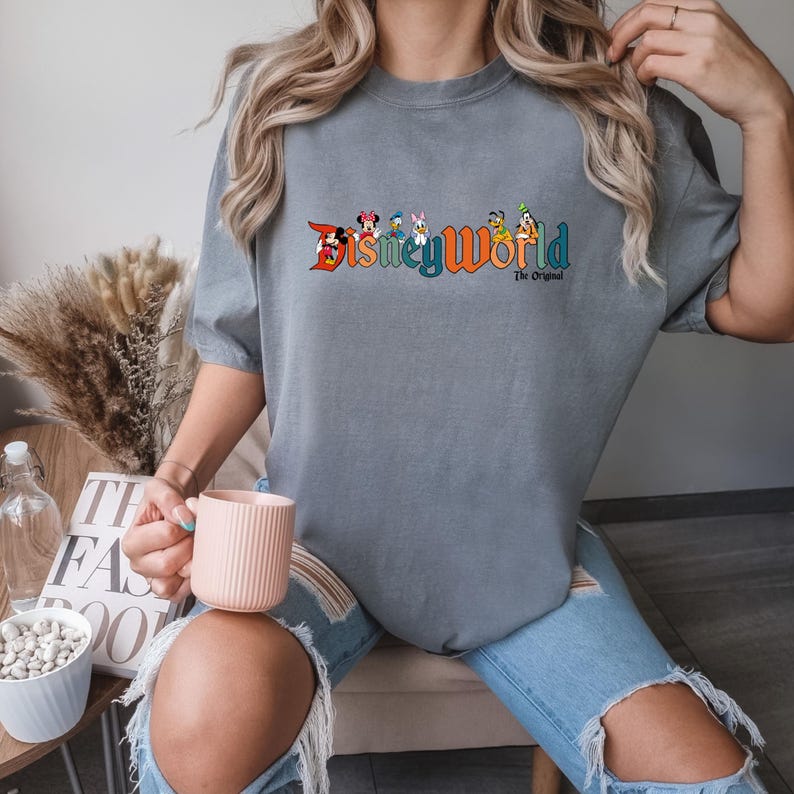 K&ouml;nnte beinhalten: Ein graues T-Shirt mit einer bunten Grafik von Disney-Charakteren und dem Text "Disneyworld The Original".