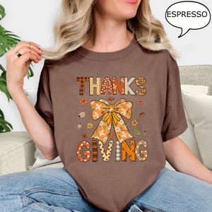 May include: A brown t-shirt with the words "THANKS GIVING" in a fall-themed design. The design includes a large orange bow, flowers, and coffee beans. The shirt is a solid brown color.