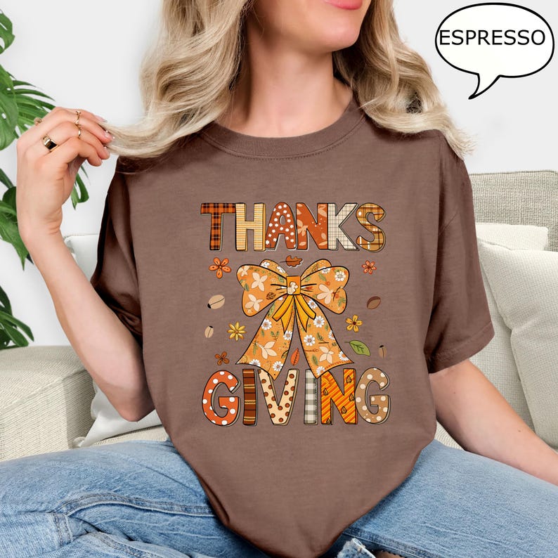 May include: A brown t-shirt with the words "THANKS GIVING" in a fall-themed design. The design includes a large orange bow, flowers, and coffee beans. The shirt is a solid brown color.