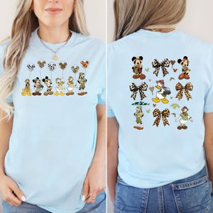 May include: Two light blue t-shirts with a safari animal theme. The front of the shirt features a line of Disney characters holding balloons. The back of the shirt features a pattern of Disney characters, balloons, and bows.