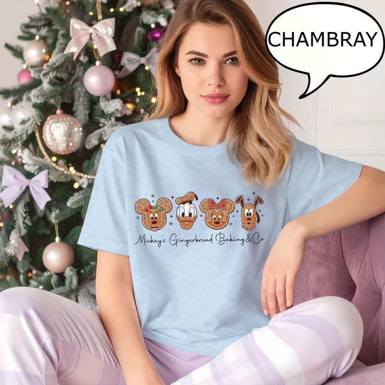 Puede incluir: Una camiseta azul claro con dise&ntilde;os de galletas de jengibre de Mickey Mouse, el Pato Donald, Minnie Mouse y Pluto. El texto "Mickey's Gingerbread Baking & Co" est&aacute; impreso debajo de las galletas. La palabra "CHAMBRAY" est&aacute; en una burbuja.