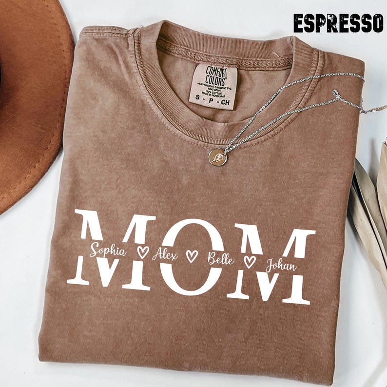 May include: Espresso-colored t-shirt with the word "MOM" in white block letters. Below the word "MOM" are the names Sophia, Alex, Belle, and Johan, each with a heart symbol. A silver necklace with a pendant is draped on the shirt.