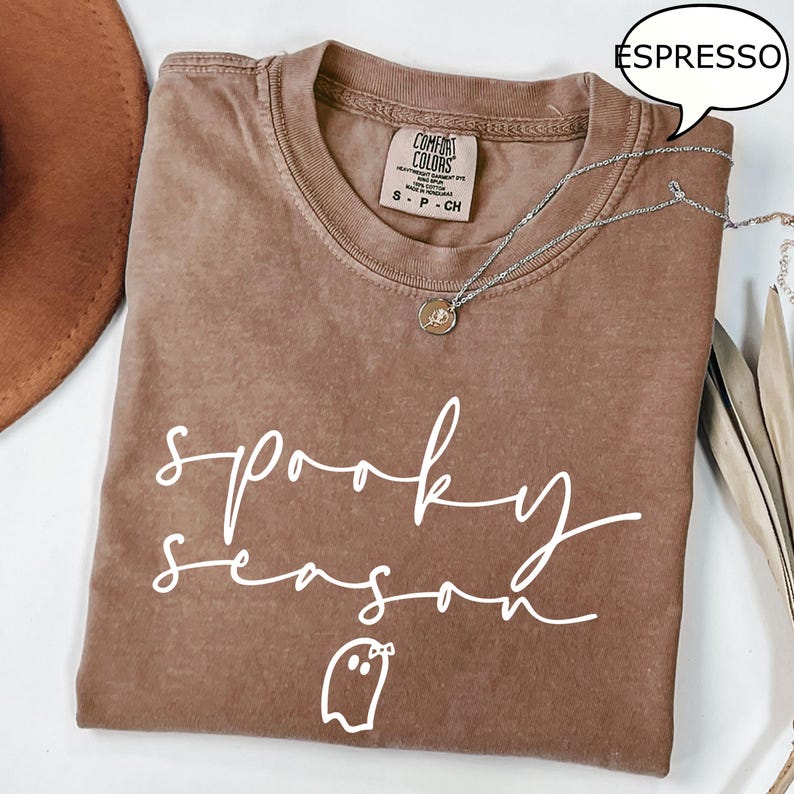May include: Espresso-coloured t-shirt featuring the words "spooky season" in white script, and a ghost graphic. The shirt has a crew neck and short sleeves. A silver necklace with a pendant is draped across the shirt.