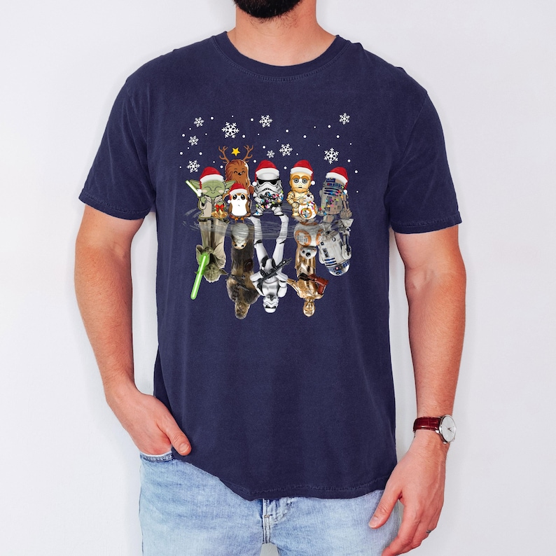 Puede incluir: Una camiseta azul marino con un gr&aacute;fico de Star Wars con tem&aacute;tica navide&ntilde;a. El gr&aacute;fico presenta personajes de la franquicia Star Wars con gorros de Pap&aacute; Noel y reflejados en el agua. Los personajes incluyen a Yoda, Chewbacca, Darth Vader, R2-D2, C-3PO y un Stormtrooper.