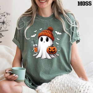 May include: A green t-shirt featuring a cartoon ghost wearing a red and white knitted hat, holding a pumpkin and a cup of orange liquid. The ghost has large eyes and a small mouth. The shirt has a festive Halloween theme.