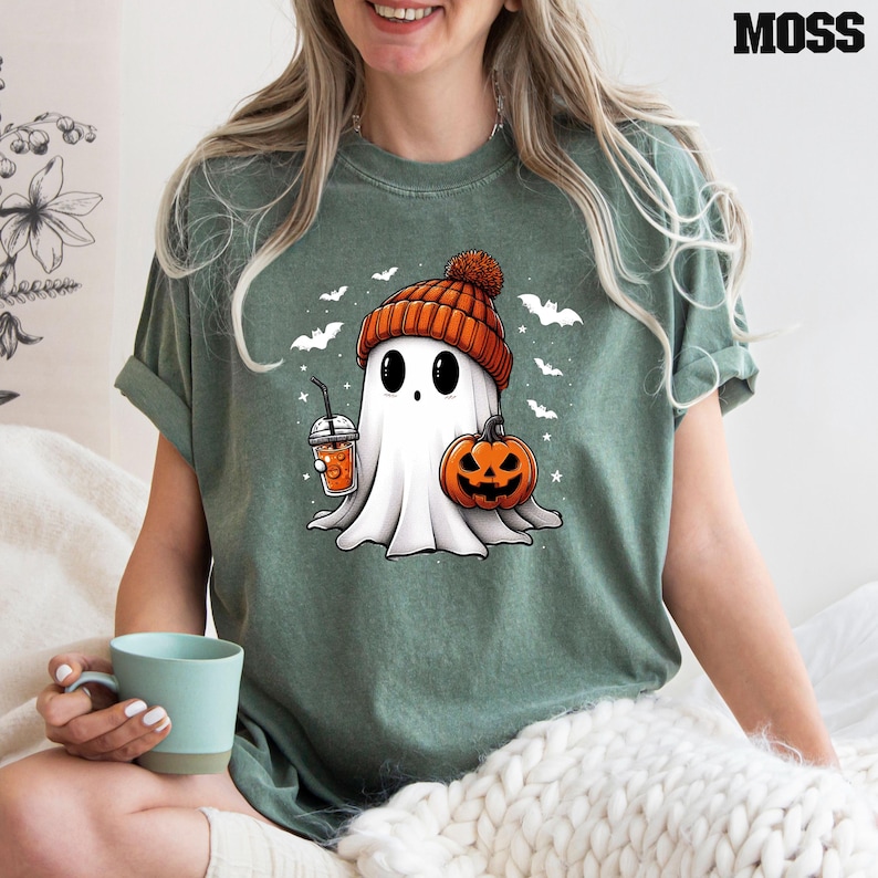 May include: A green t-shirt featuring a cartoon ghost wearing a red and white knitted hat, holding a pumpkin and a cup of orange liquid. The ghost has large eyes and a small mouth. The shirt has a festive Halloween theme.