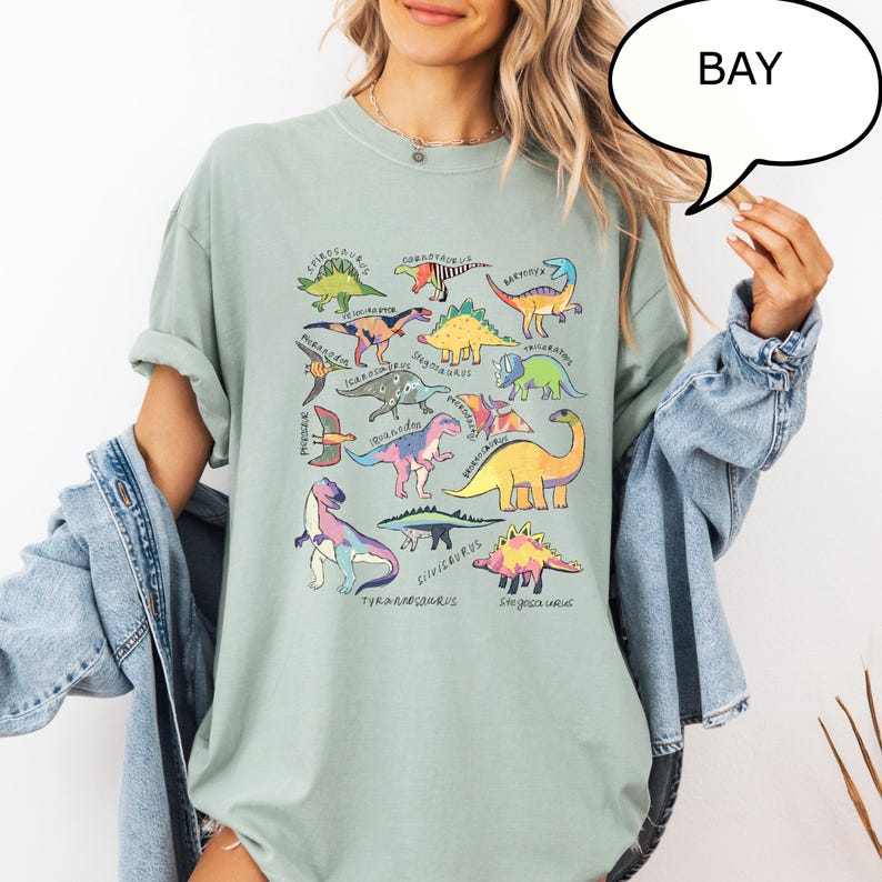 May include: Sage green t-shirt featuring a colorful dinosaur graphic. The graphic includes various dinosaur illustrations with their names, such as Tyrannosaurus and Stegosaurus. The word "BAY" is in a speech bubble.