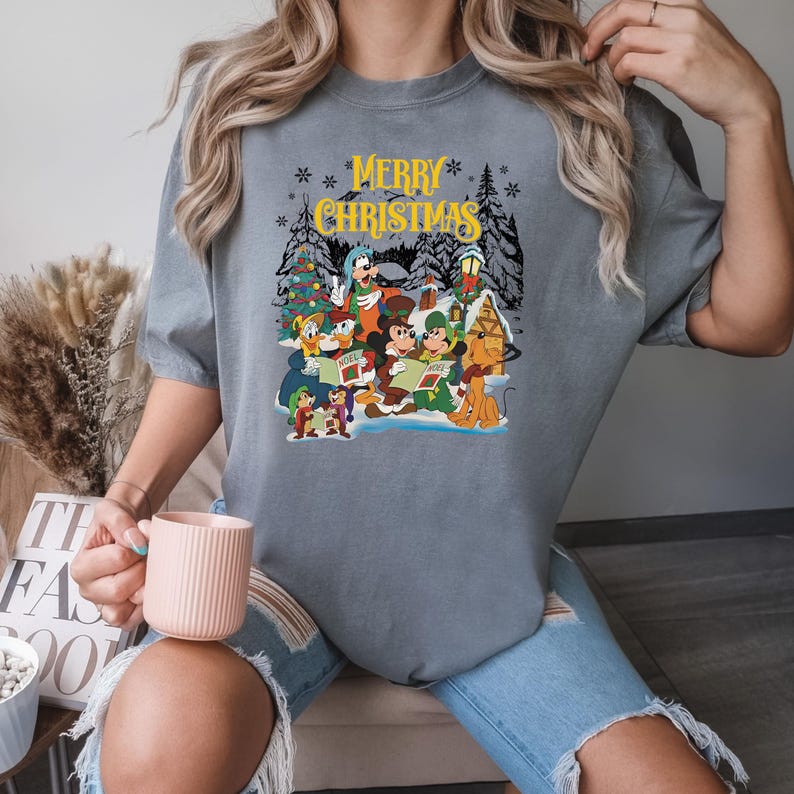 Puede incluir: Una camiseta gris con un gr&aacute;fico navide&ntilde;o con Mickey Mouse y sus amigos cantando villancicos. El texto "Merry Christmas" est&aacute; impreso en amarillo en la camiseta.