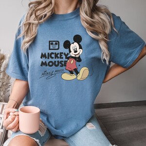 May include: A blue t-shirt with a vintage Mickey Mouse graphic. The graphic features Mickey Mouse in his signature red shorts and yellow shoes. The text "Mickey Mouse" is printed in white below the graphic.