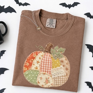 May include: A brown crewneck t-shirt featuring a large, central pumpkin appliqué made from various fabric patches, including floral, gingham, and lace patterns. The shirt has a Comfort Colors brand label at the inner neckline and a silver necklace.