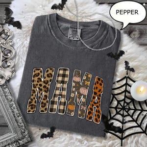 May include: A dark gray Comfort Colors t-shirt with the word "MAMA" in a mix of leopard print, plaid, floral, and pumpkin patterns. The shirt is decorated with Halloween-themed accents, including bats and a spiderweb.