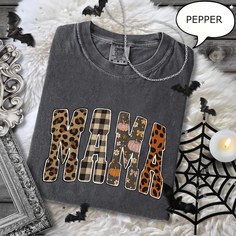May include: A dark gray Comfort Colors t-shirt with the word "MAMA" in a mix of leopard print, plaid, floral, and pumpkin patterns. The shirt is decorated with Halloween-themed accents, including bats and a spiderweb.