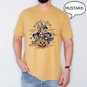 May include: Mustard-colored t-shirt featuring a Halloween-themed graphic of Disney characters, including Mickey Mouse, Minnie Mouse, Donald Duck, and Goofy, surrounding a carved pumpkin. The word "MUSTARD" is in a speech bubble.