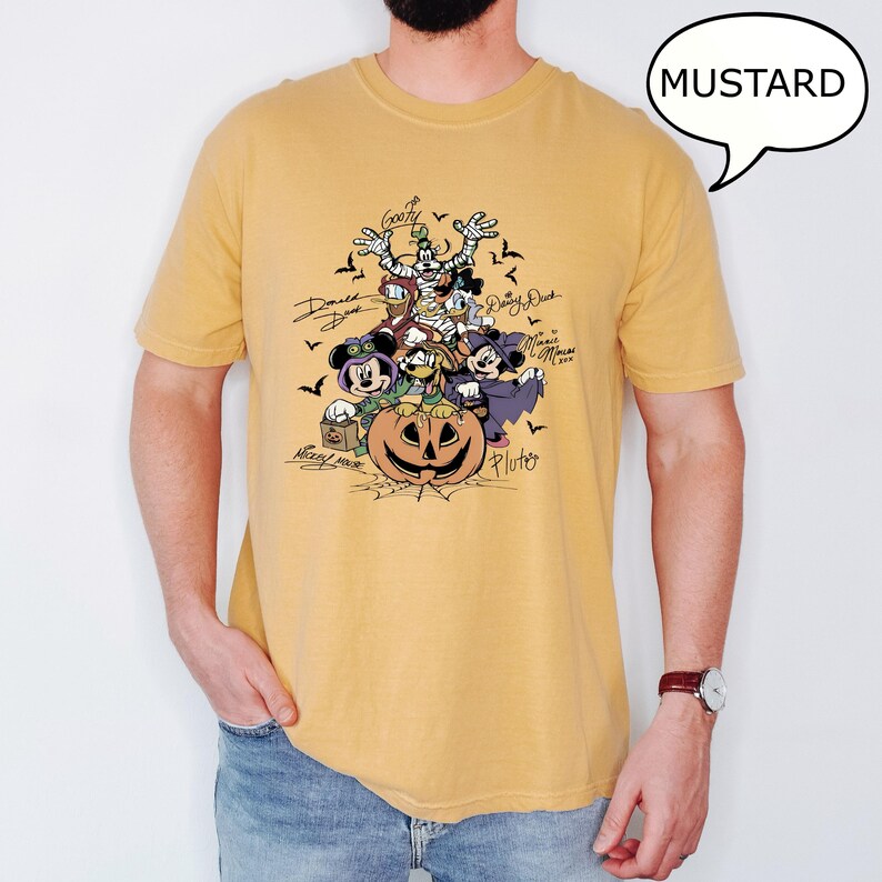 May include: Mustard-colored t-shirt featuring a Halloween-themed graphic of Disney characters, including Mickey Mouse, Minnie Mouse, Donald Duck, and Goofy, surrounding a carved pumpkin. The word "MUSTARD" is in a speech bubble.