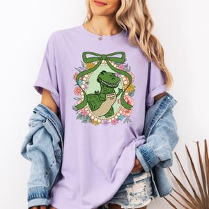 May include: A purple t-shirt with a cartoon image of a green dinosaur wearing a bow. The dinosaur is surrounded by flowers and pearls.