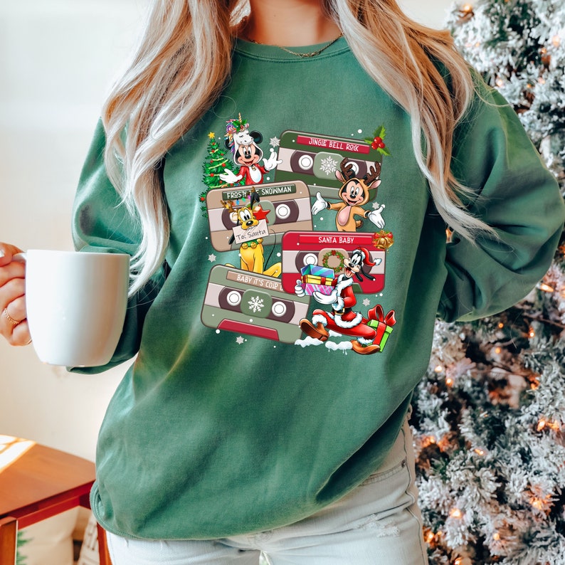 Puede incluir: Sudadera verde con un dise&ntilde;o navide&ntilde;o con personajes de dibujos animados y cintas de casete. Las cintas tienen t&iacute;tulos como "Jingle Bell Rock", "Frosty", "Snowman", "Santa Baby" y "Baby It's Cold Outside".