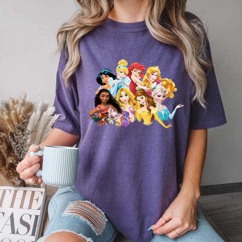 Puede incluir: Camiseta morada con un gr&aacute;fico de princesas Disney, incluyendo Ariel, Bella, Cenicienta, Elsa, Jasmine, Moana, Mulan, Pocahontas, Rapunzel y Blancanieves.