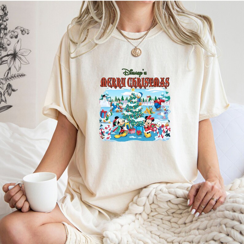 May include: A cream colored t-shirt with a Christmas themed graphic featuring Mickey Mouse and other Disney characters. The graphic says "Disney's Merry Christmas".