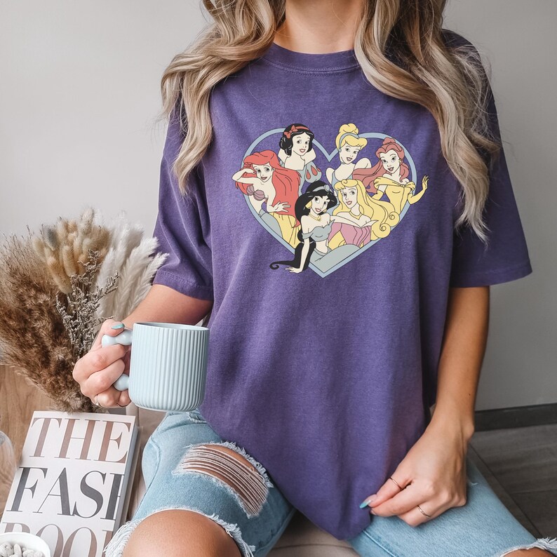 Pu&ograve; includere: Maglietta viola con una grafica a forma di cuore di principesse Disney. Le principesse sono disposte a forma di cuore e includono Biancaneve, Cenerentola, Ariel, Belle, Jasmine e Aurora.