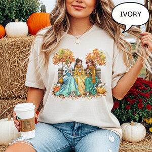 May include: Ivory-colored t-shirt featuring a graphic of three princesses sitting on a bench, surrounded by autumn leaves and pumpkins. The princesses are wearing dresses and scarves, holding cups. The word "IVORY" is in a speech bubble.