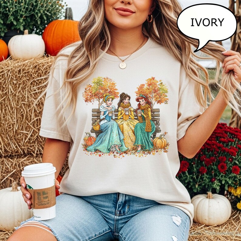 May include: Ivory-colored t-shirt featuring a graphic of three princesses sitting on a bench, surrounded by autumn leaves and pumpkins. The princesses are wearing dresses and scarves, holding cups. The word "IVORY" is in a speech bubble.