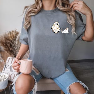May include: A grey t-shirt featuring a graphic of a ghost wearing a patterned sheet and walking a dog ghost. The dog ghost is wearing a white sheet.