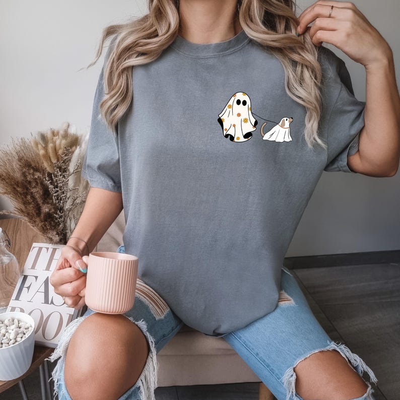 May include: A grey t-shirt featuring a graphic of a ghost wearing a patterned sheet and walking a dog ghost. The dog ghost is wearing a white sheet.