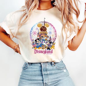 May include: A white t-shirt with a colorful graphic of Mickey Mouse and other Disney characters celebrating Disneyland's 70th anniversary. The graphic includes fireworks, a castle, and the text "Disneyland Resort".