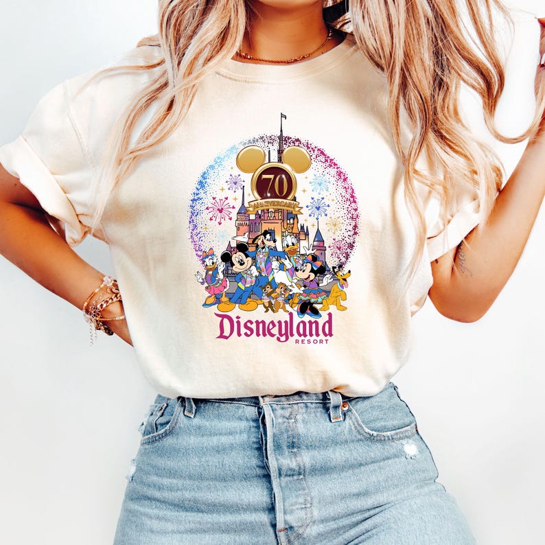 May include: A white t-shirt with a colorful graphic of Mickey Mouse and other Disney characters celebrating Disneyland's 70th anniversary. The graphic includes fireworks, a castle, and the text "Disneyland Resort".