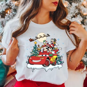 May include: White t-shirt with a Christmas-themed design featuring Mickey Mouse, Donald Duck, Goofy, and Pluto riding in a red Lightning McQueen car. Santa Claus is flying in the sky above them.