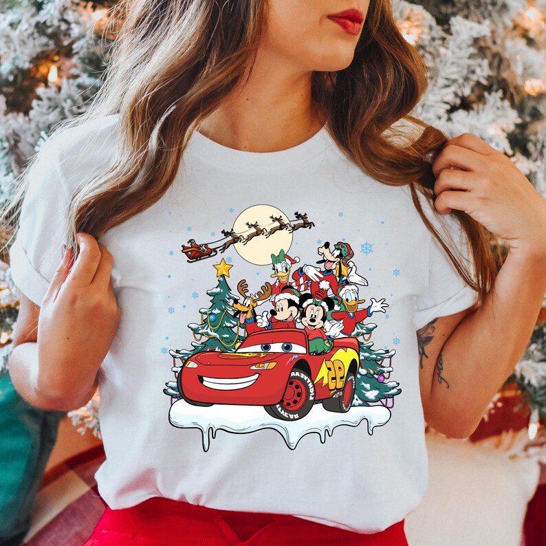 May include: White t-shirt with a Christmas-themed design featuring Mickey Mouse, Donald Duck, Goofy, and Pluto riding in a red Lightning McQueen car. Santa Claus is flying in the sky above them.