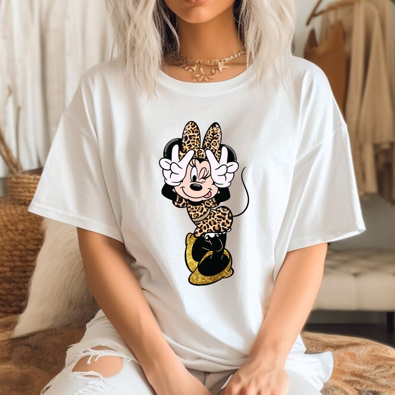K&ouml;nnte beinhalten: Wei&szlig;es T-Shirt mit einer Cartoon-Figur von Minnie Mouse in einem Leopardenmuster-Outfit. Minnie macht mit ihren H&auml;nden ein Friedenszeichen. Das Design umfasst die Farben Schwarz, Gold und Braun.