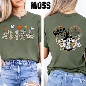 Puede incluir: Camiseta verde musgo con un dise&ntilde;o de Halloween. La parte delantera muestra personajes de dibujos animados momificados sosteniendo globos. La parte trasera muestra la palabra "Boo!!" con una telara&ntilde;a y personajes. La palabra "MOSS" est&aacute; impresa arriba.