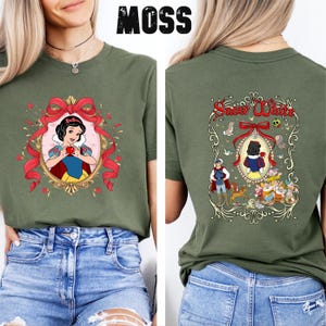 May include: Olive green t-shirt with a Snow White graphic on the front and back. The front features Snow White holding an apple in a gold frame with a red bow. The back has a Snow White design with the words "Snow White".