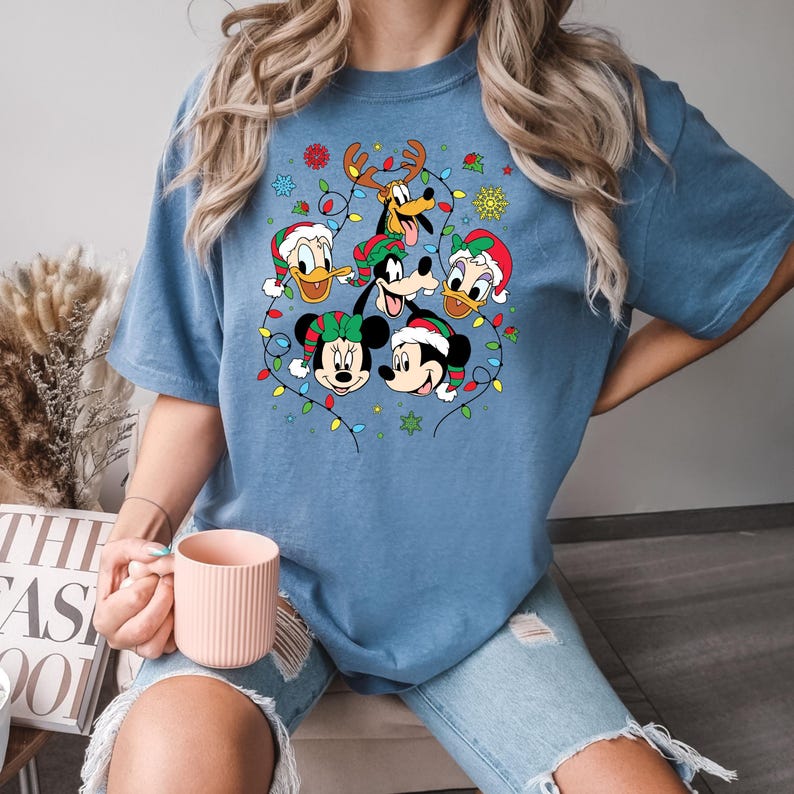 May include: A blue t-shirt with a Christmas-themed design featuring Mickey Mouse, Minnie Mouse, Donald Duck, Goofy, and Pluto wearing Santa hats and surrounded by Christmas lights.
