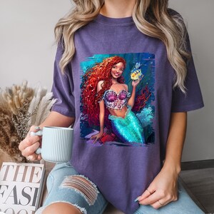 May include: Purple t-shirt featuring a graphic print of a red-haired mermaid with a seashell bra and a turquoise tail. The mermaid is holding a yellow fish.
