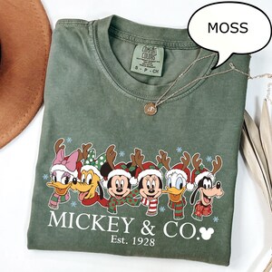 Puede incluir: Una camiseta verde musgo con un dise&ntilde;o de personajes de Disney con tem&aacute;tica navide&ntilde;a. La camiseta tiene el texto "Mickey & Co. Est. 1928". Un collar plateado con un colgante est&aacute; sobre la camiseta.