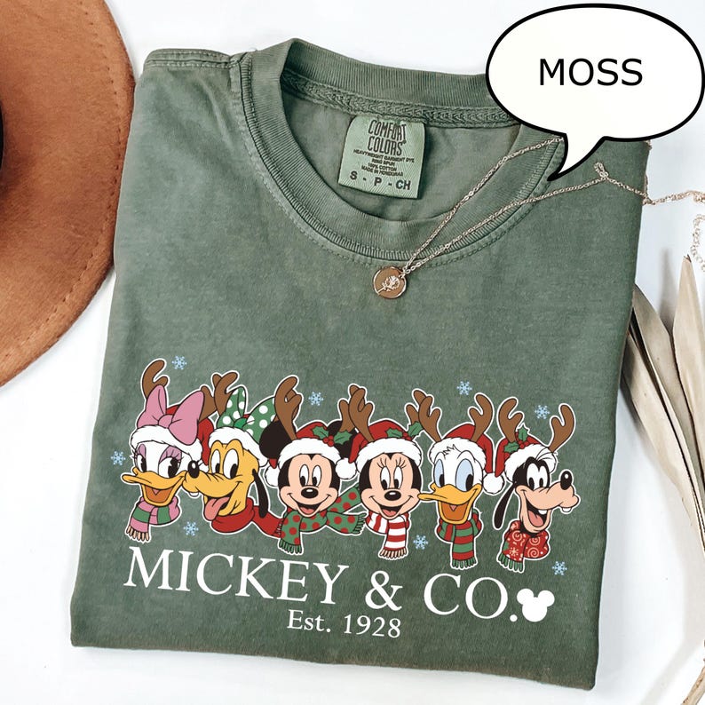 Puede incluir: Una camiseta verde musgo con un dise&ntilde;o de personajes de Disney con tem&aacute;tica navide&ntilde;a. La camiseta tiene el texto "Mickey & Co. Est. 1928". Un collar plateado con un colgante est&aacute; sobre la camiseta.
