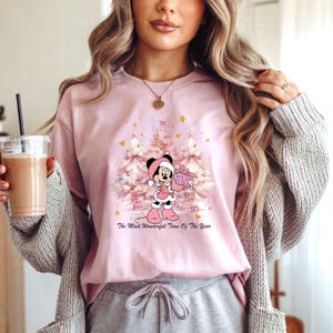 May include: A light pink t-shirt featuring a graphic of Minnie Mouse in a winter outfit holding a gift in front of Christmas trees. The text "The Most Wonderful Time Of The Year" is below the graphic.