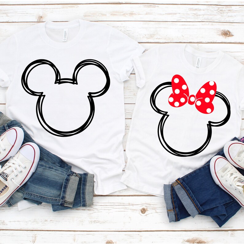 May include: Two white t-shirts with black outlines of Mickey Mouse's head. One shirt has a red bow with white polka dots on top of the head. The shirts are laid out on a white wooden surface with blue jeans and white sneakers.