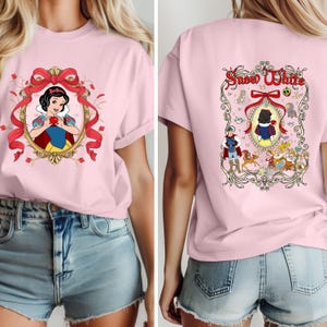 May include: A light pink t-shirt featuring a Snow White design. The front of the shirt displays Snow White holding an apple within a decorative frame. The back of the shirt has the words "Snow White" and a scene with characters.