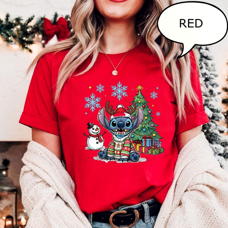 Puede incluir: Camiseta roja con un personaje de dibujos animados con un gorro de reno y un jersey navide&ntilde;o, con un mu&ntilde;eco de nieve, un &aacute;rbol de Navidad y copos de nieve. La palabra "RED" est&aacute; en una burbuja.