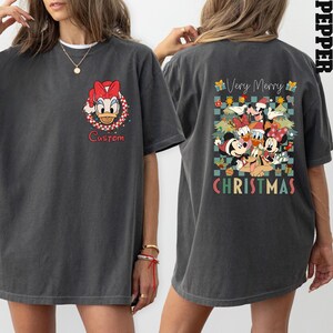 Pode incluir: Camiseta cinza escuro com um desenho de Margarida com um la&ccedil;o vermelho e a palavra "Custom". A parte de tr&aacute;s da camiseta exibe um design de Natal com Mickey, Minnie, Donald e Pateta, com as palavras "Very Merry Christmas".