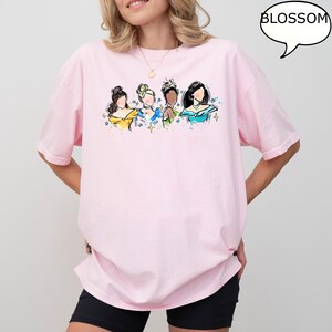 May include: A light pink t-shirt featuring a watercolor illustration of four women with various hairstyles and colorful dresses. The word "BLOSSOM" is in a speech bubble in the upper right corner. The shirt is a relaxed fit.