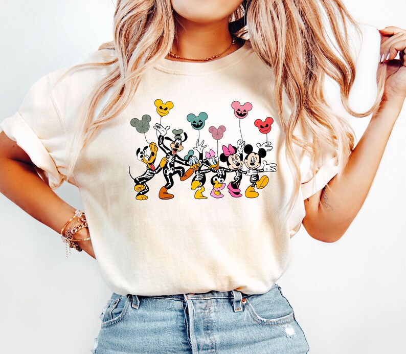 May include: A white t-shirt with a graphic of Mickey Mouse, Minnie Mouse, Donald Duck, Goofy, and Pluto dressed in skeleton costumes holding colorful balloons. The balloons are shaped like Mickey Mouse ears and have a Halloween theme.