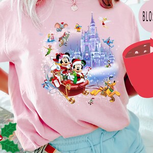 Puede incluir: Camiseta rosa con un dise&ntilde;o navide&ntilde;o con Mickey Mouse y Minnie Mouse en un trineo tirado por Pluto frente al Castillo de Cenicienta. El dise&ntilde;o tambi&eacute;n incluye otros personajes de Disney como Campanilla, Aladdin y Jasmine.