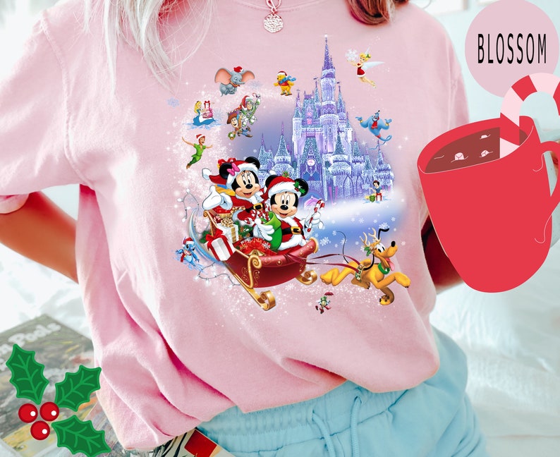 Puede incluir: Camiseta rosa con un dise&ntilde;o navide&ntilde;o con Mickey Mouse y Minnie Mouse en un trineo tirado por Pluto frente al Castillo de Cenicienta. El dise&ntilde;o tambi&eacute;n incluye otros personajes de Disney como Campanilla, Aladdin y Jasmine.