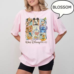 May include: A light pink t-shirt featuring a grid of Disney characters, including Mickey Mouse, Donald Duck, and Stitch. The words "Walt Disney World" are printed below the grid in a cursive font. A speech bubble with the word "BLOSSOM" is in the top right corner.